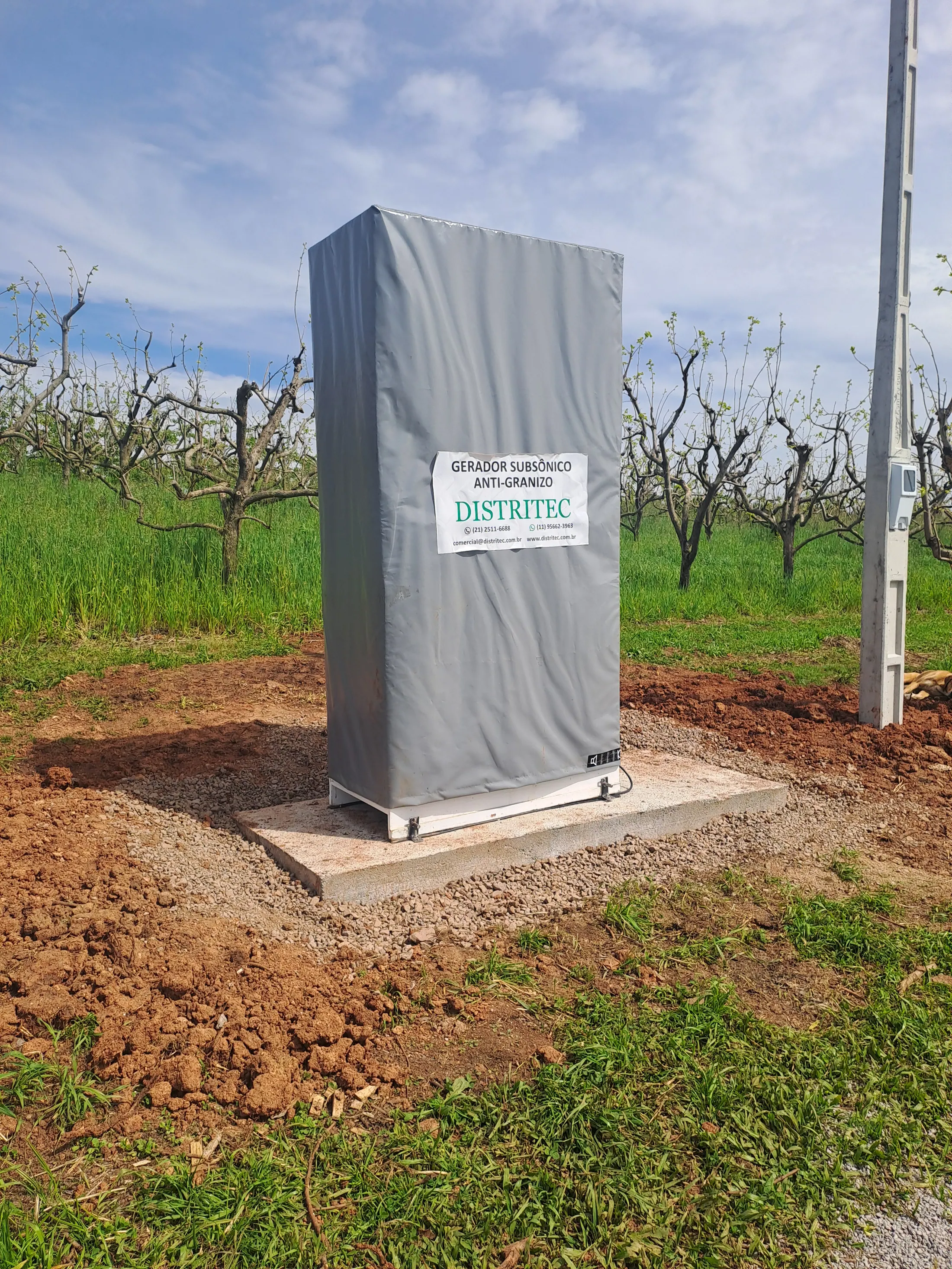 Gerador subsônico DGS-AG com cobertura cinza sobre uma base de concreto, em um pomar