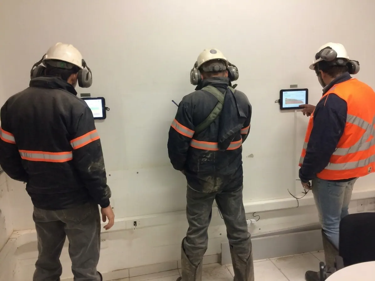 Três trabalhadores de capacete diante de um tablet fixado na parede, para o teste de alerta