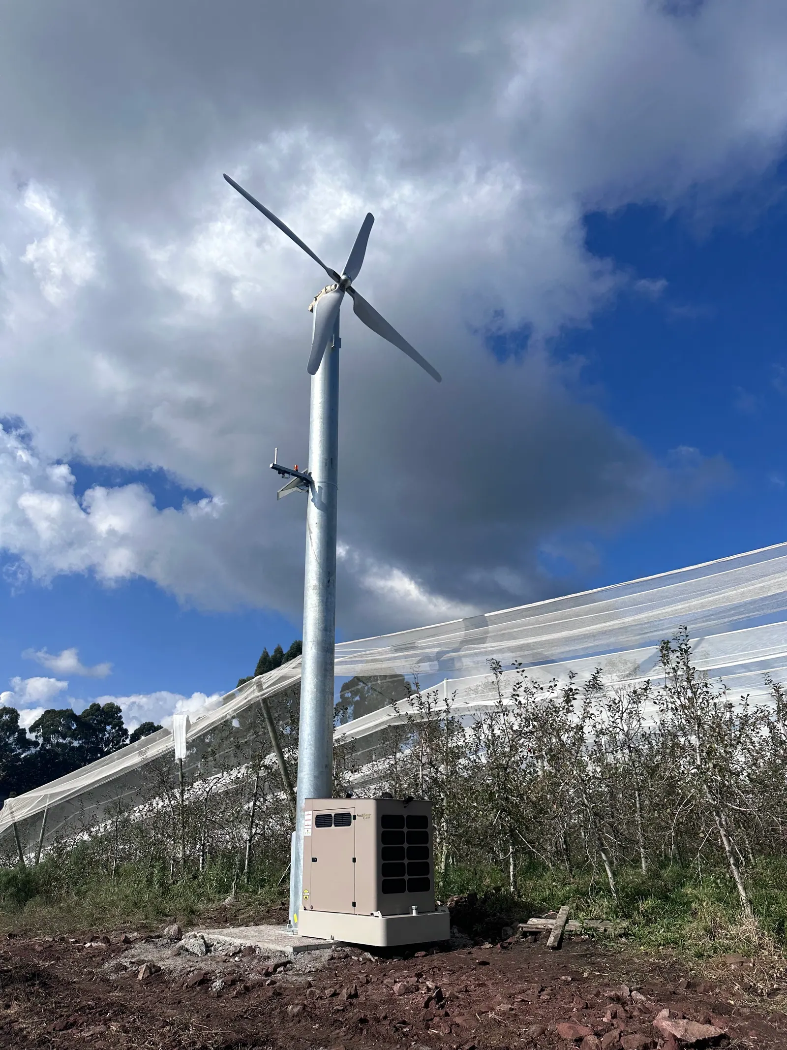 Ventilador anti-geada fixo instalado junto à tela anti-granizo de um pomar